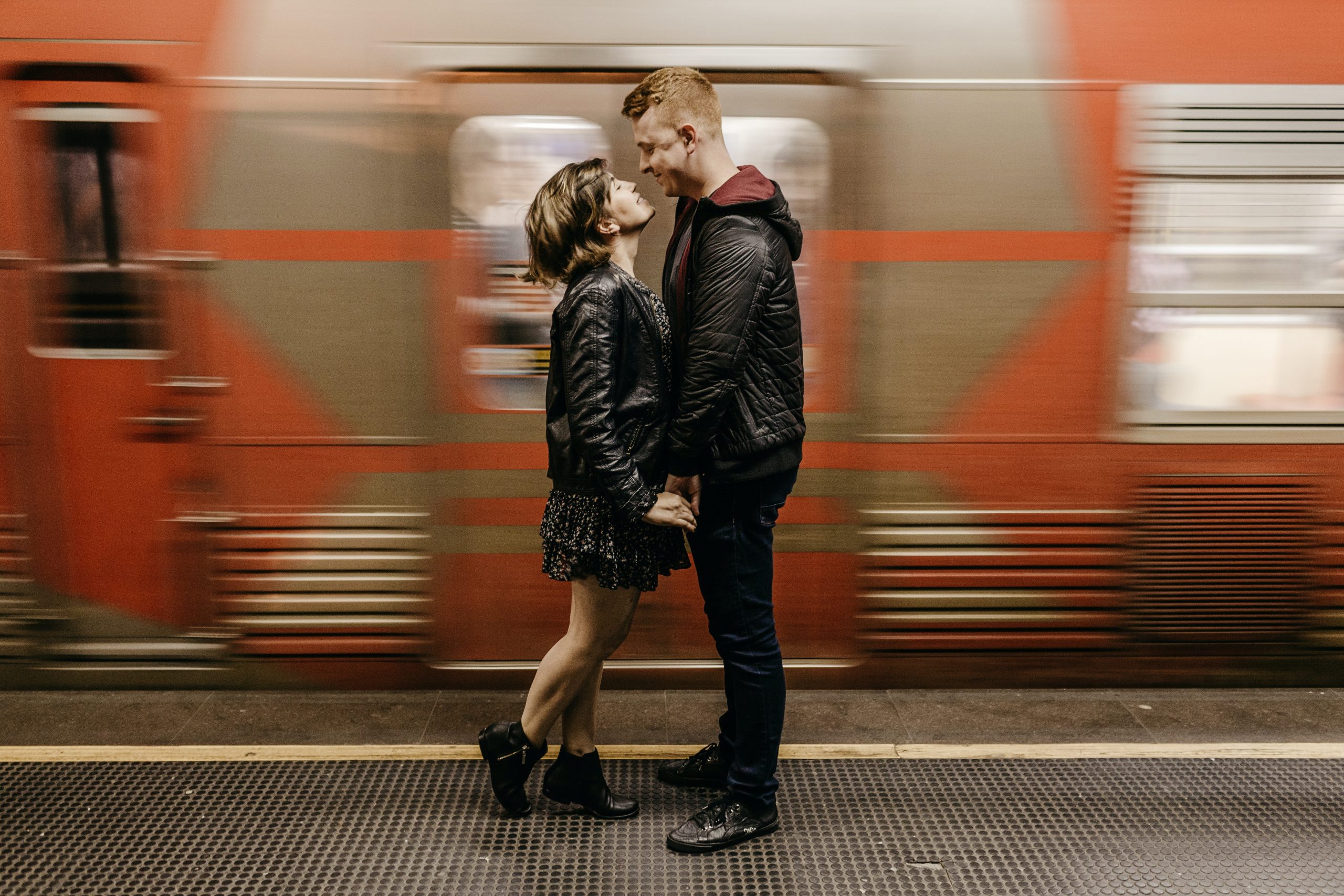 hoto of Smiling Couple Standing on Train Station Platform While Holding Hands as a Train Passes